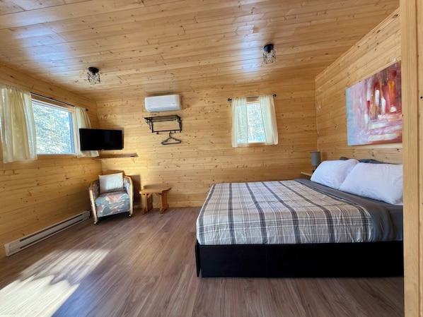 A wooden cabin room with a bed, a chair, a small table, and a mounted TV.