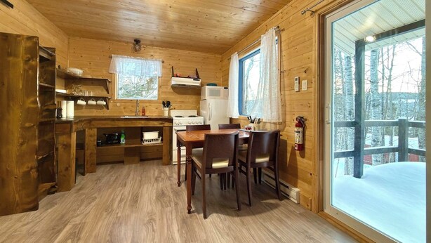 A wooden cabin interior with a dining table, chairs, a stove, and a large window.