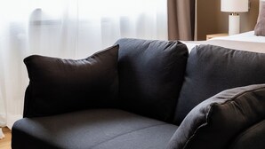 A modern living room with a dark sofa, a wooden floor, a white lamp, and a view through sheer curtains. - Charming Studio | Downtown | 2min Metro | 401 (BRUXELLES)