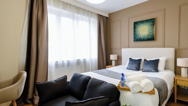 A hotel room with a bed, a sofa, a bedside table with a lamp, a window with curtains, and a wall-mounted artwork. - Charming Studio | Downtown | 2min Metro | 401 (BRUXELLES)