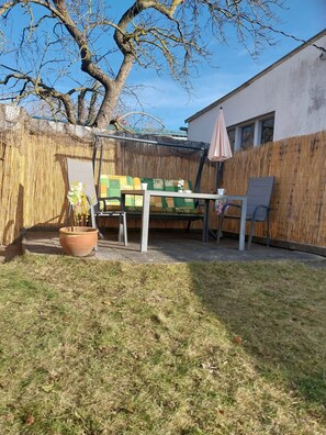 Un patio trasero con área de terraza, un árbol grande y una valla de bambú.