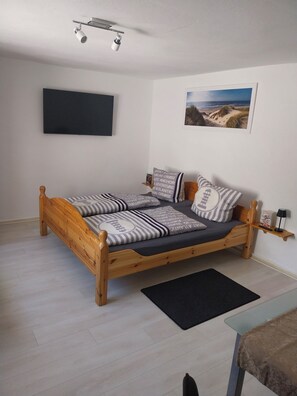 Una cama de madera con ropa de cama a rayas, un televisor negro montado en la pared, un cuadro con un paisaje costero enmarco, y una mesita de noche pequeña con una lámpara.