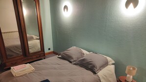 Een opgemaakt bed met een gevouwen handdoek en een plaid, een houten nachtkastje met een lamp, en een grote spiegel aan de muur.