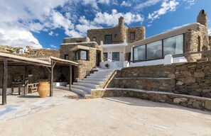 A stone building with a staircase leading to a white upper level, a large window, and a covered outdoor area with a table and chairs. - ⋆ 5BR Oceanside View Near Lighthouse- Pharos VM ⋆ (Faros Armenistis)