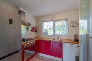 Une cuisine avec des meubles de couleur rouge, un lave-vaisselle Bosch, une cuisinière et une fenêtre avec des stores.