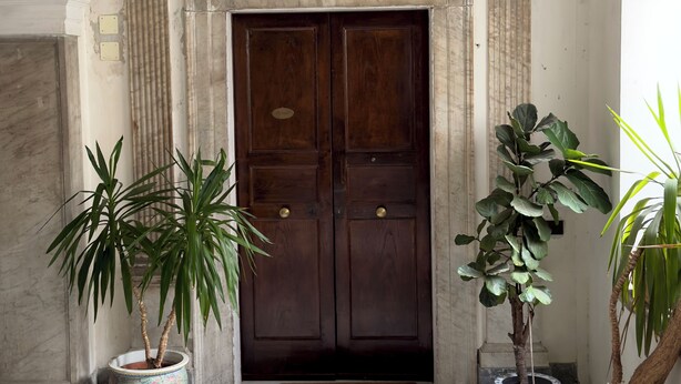 A grand entrance with a dark wooden door, flanked by ornate columns and a decorative ceiling. There are potted plants on either side of the door. - Palazzo Carafa Santangelo - historic residence of the 400'in the heart of the ancient city (Napoli)