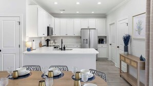 A modern kitchen with white cabinetry, a central island, and a dining area with a wooden table set for four. - New Crescent Beach luxury home w/pool & NO pet fee (North Myrtle Beach)