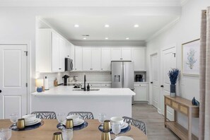 A modern kitchen with white cabinetry, a central island, and a dining area with a wooden table set for four. - New Crescent Beach luxury home w/pool & NO pet fee (North Myrtle Beach)