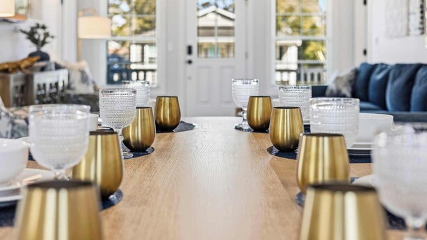A dining table set with glassware and gold-colored candle holders in a well-lit room with a view of a building through a window. - New Crescent Beach luxury home w/pool & NO pet fee (North Myrtle Beach)