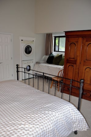 Une chambre à coucher comprenant un lit, un lave-linge, une armoire en bois, une fenêtre avec des rideaux et une chaise.