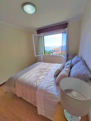 Une chambre à coucher avec un lit, une lampe de chevet, une fenêtre avec des rideaux et une vue sur des immeubles et la mer.