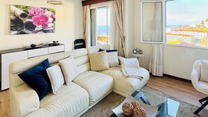 Un salon moderne avec un canapé d’angle beige, une table basse en verre et une grande fenêtre donnant sur la mer et les bâtiments.