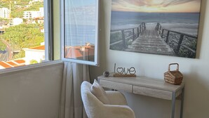 Une pièce avec vue sur un paysage côtier, visible par une fenêtre, comprenant un bureau avec un panier en osier, une chaise et un tableau accroché au mur représentant une jetée.