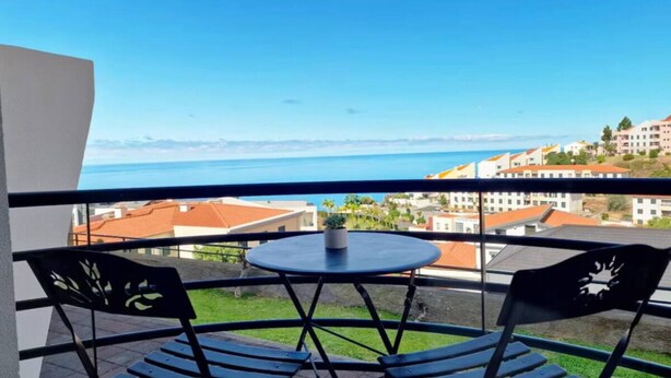 Un balcon avec une table et des chaises, donnant sur une ville côtière avec ses bâtiments et un ciel dégagé.