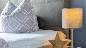 Ein Bett mit grauem Kopfteil, einem weißen Kissen und einem gemusterten Kissen. Ein runder Nachttisch mit einer Lampe und einem sternförmigen Gegenstand darauf.