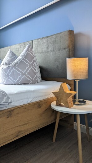 A bed with a grey headboard, a white pillow, and a patterned pillow. There is a round wooden nightstand with a lamp and a star-shaped object on it. - Comfortable holiday apartment for 2–3 guests near old town with parking & WiFi (Wernigerode)