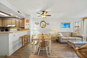A spacious living area with a dining table, sofa, and a kitchen area with wooden cabinets and a built-in oven. - Pelican's Watch 211- 3 Bedroom- Outdoor Pool (Myrtle Beach)