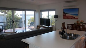 Cozinha moderna com ilha integrada, área de jantar com mesa e cadeiras, uma janela grande com vista para o verde e uma TV de tela plana em um suporte.