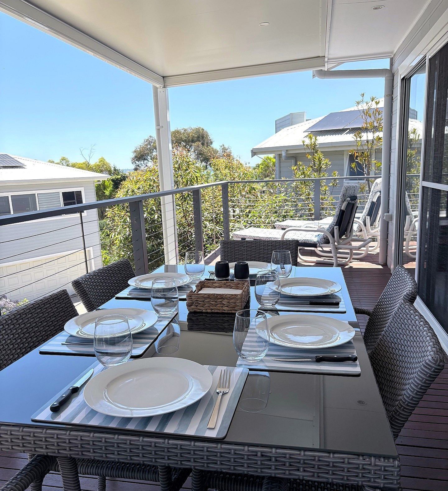An outdoor dining area with a table set for four, surrounded by wicker chairs, on a balcony with a view of a residential area.
