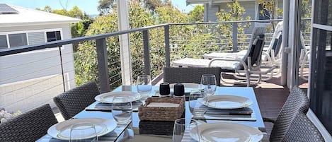 An outdoor dining area with a table set for four, surrounded by wicker chairs, on a balcony with a view of a residential area.