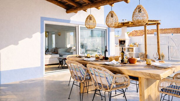 Un área de comedor al aire libre con sillas de mimbre, una mesa de madera dispuesta para una comida y luces colgantes de mimbre.