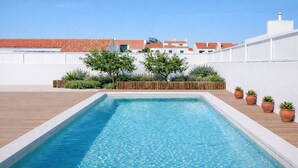 Una piscina rectangular con un muelle de madera que la rodea, frente a un muro blanco con una fila de macetas y una cerca de madera.