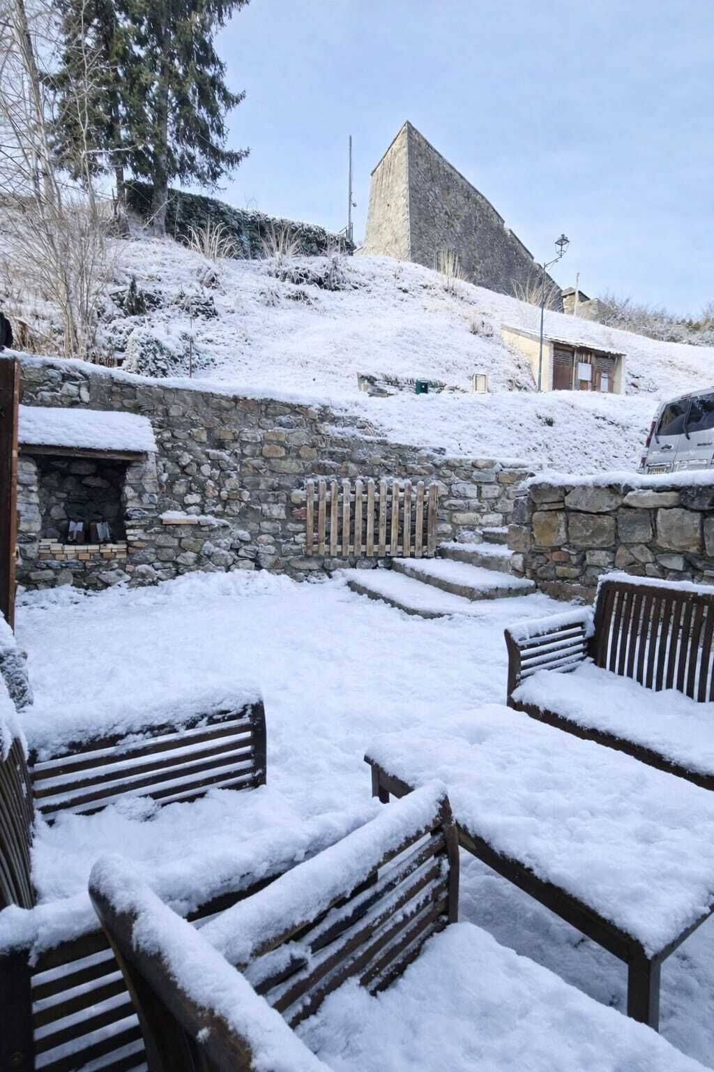 A snow-covered outdoor area with stone walls, wooden benches, and a stone building in the background.