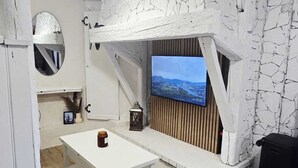 Una habitación acogedora con una mesa blanca, una lámpara colgante, un televisor fijado a una pared de piedra y piso de madera.