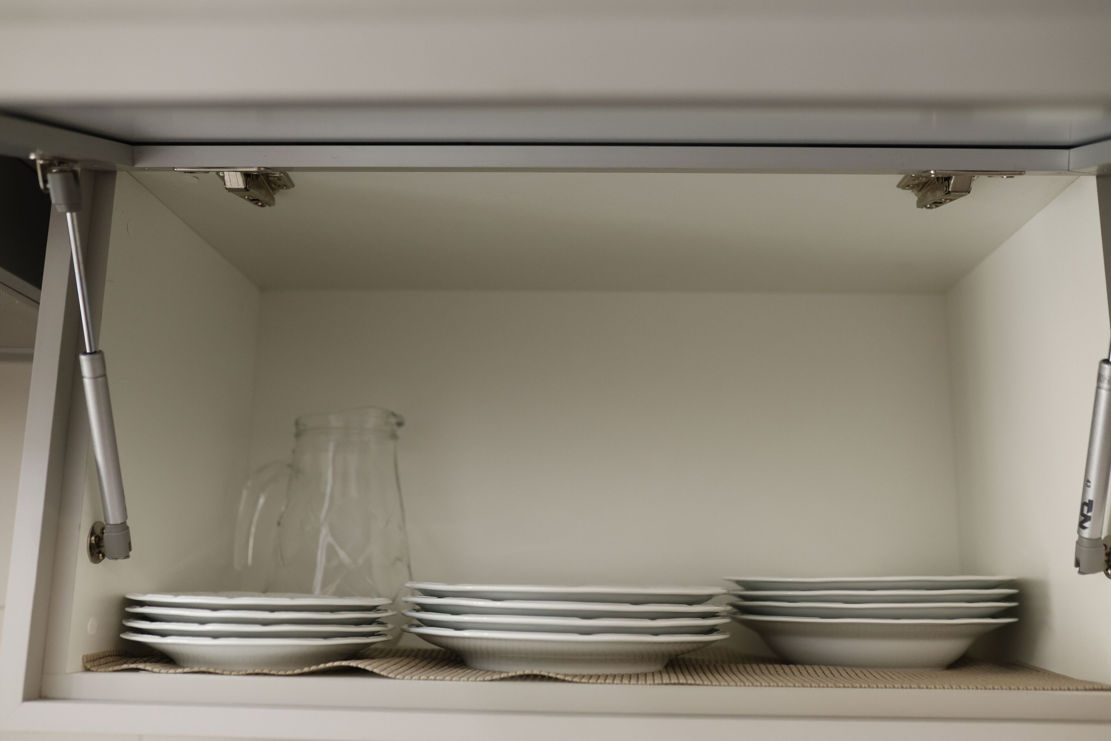 Ein Küchenschrank mit weißem Innenraum, der stapelweise weiße Teller, eine klare Glasvase und eine gewebte Tischmatte beherbergt.