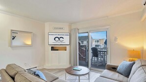 A modern living room with a sofa, a coffee table, and a sliding glass door leading to a balcony. A sign on the wall reads "BEACH HOUSE" and "OCEAN CITY VACATION HOMES." - Harbour Bay 5, Beautiful 3BR Modern Bayside Townhome, Quiet! (Ocean City)