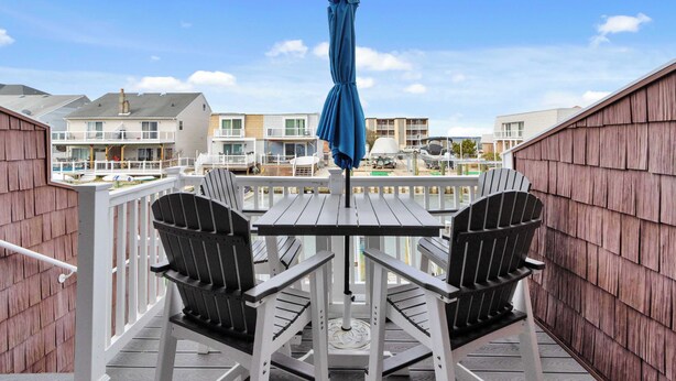 A balcony with a table and two chairs, overlooking a residential area with houses and a view of the water. - Harbour Bay 5, Beautiful 3BR Modern Bayside Townhome, Quiet! (Ocean City)