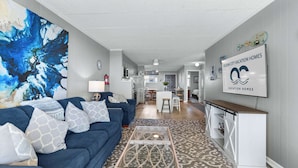 Uma sala de estar moderna com um sofá azul, uma mesa de centro e uma grande pintura abstrata. Uma área de cozinha é visível ao fundo, com um aviso da Ocean City Vacation Homes.