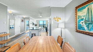 Uma ampla área de jantar com uma mesa de madeira e cadeiras de vime, uma cozinha ao fundo, e um quadro grande na parede.