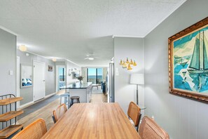 Un vaste espace dédié à la salle à manger, avec une table en bois et des chaises en osier, une cuisine à l’arrière-plan et un grand tableau accroché au mur.