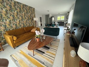 A modern living room with a sofa, coffee table, and a TV. There is a kitchen in the background with a dining area. - "LE FAIRWAY - Single-family home, single-story, 96 m², 5 minutes from Troyes (Bréviandes)