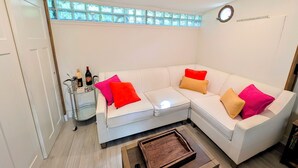 Een witte hoekbank met kleurrijke kussens, een klein bijzettafeltje met een fles en een houten salontafel.