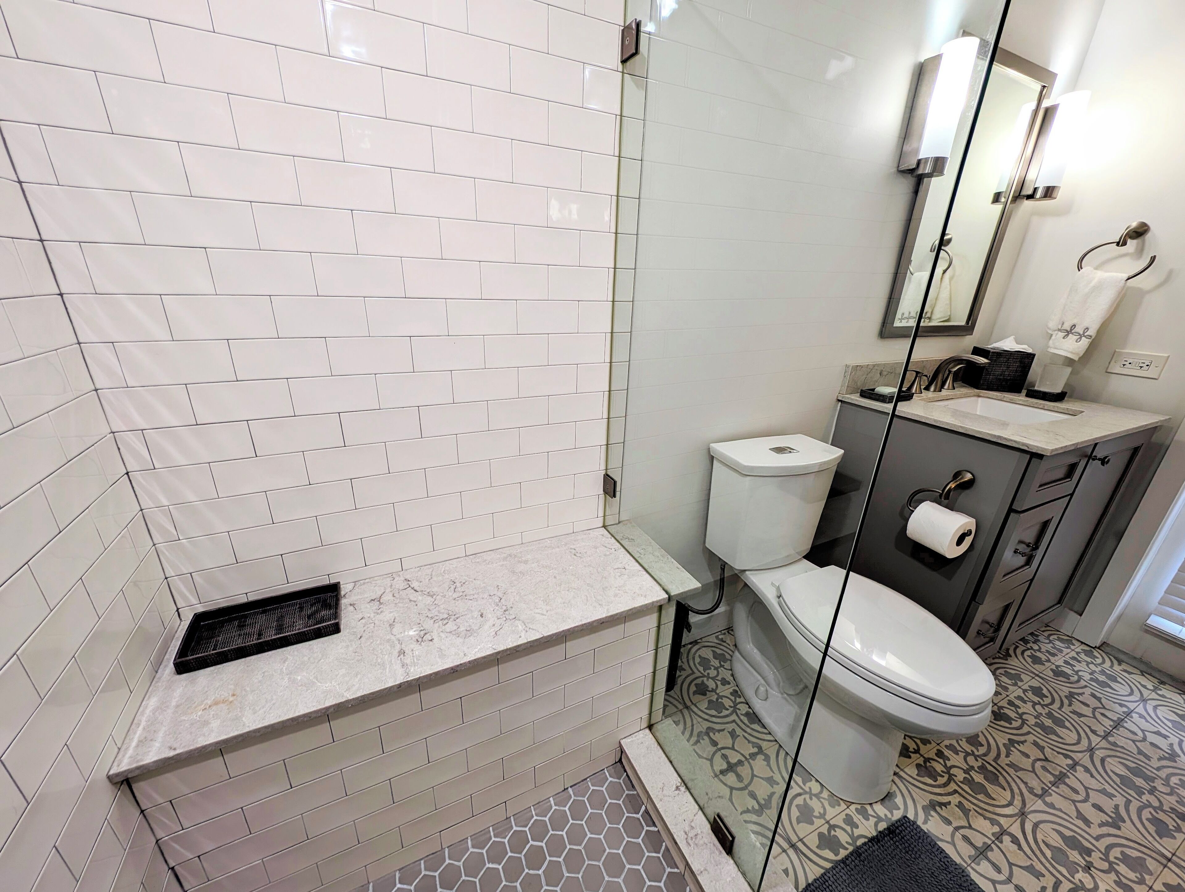 Ein modernes Badezimmer mit weißen, rechteckigen Fliesen, einer Marmorarbeitsplatte, einer Toilette, einem Waschbecken mit Spiegel und Fliesenboden mit Muster.