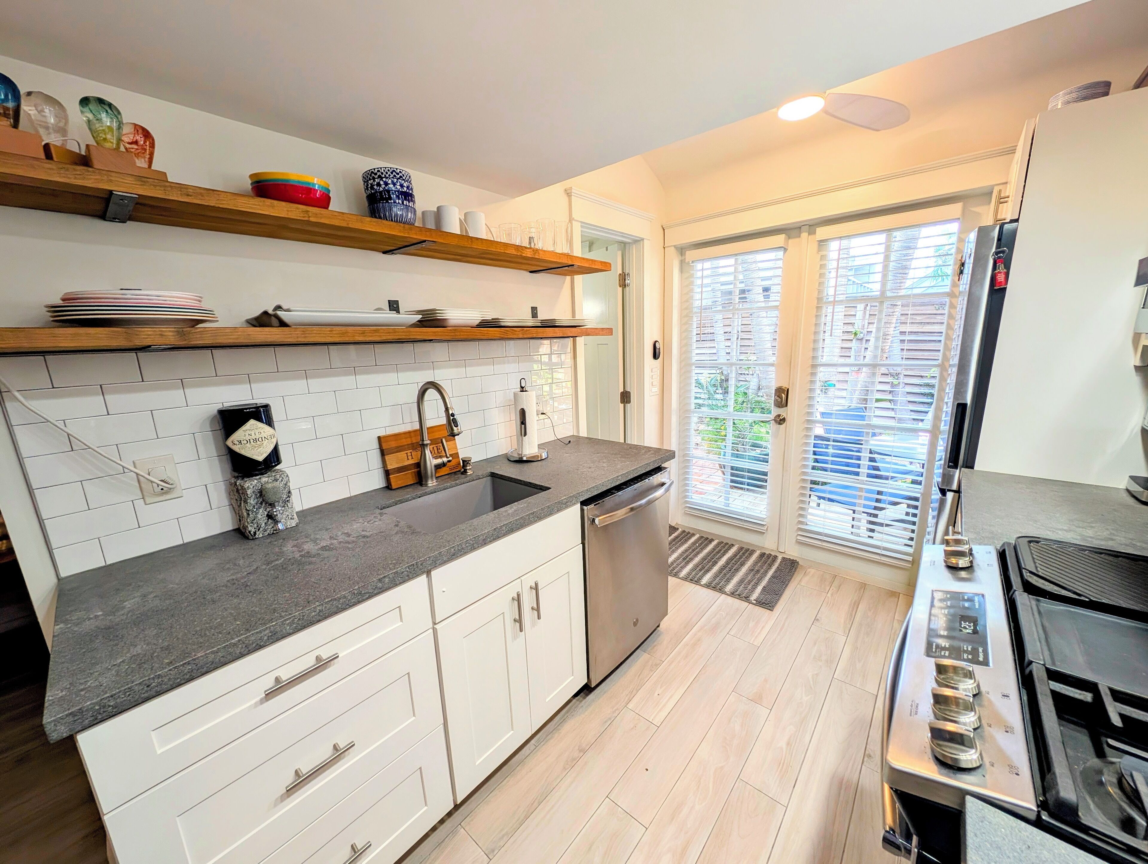 A modern kitchen with white cabinets, a granite countertop, and a stainless steel sink. There are wooden shelves above the sink holding dishes and a decorative vase. A black stove is visible on the right side.
