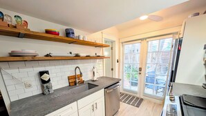 Een moderne keuken met witte kasten, een granieten aanrecht en een roestvrijstalen spoelbak. Boven de spoelbak staan houten planken met servies en een decoratieve vaas. Aan de rechterkant is een zwarte kookplaat zichtbaar.