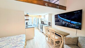 Een compacte leefruimte met een kitchenette, een eethoek met stoelen, een bank en een televisie aan de muur waarop een trein te zien is.