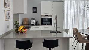 Una cucina moderna con mobili bianchi, forno integrato e macchina per il caffè. Sul piano di lavoro c'è una ciotola di frutta e un'area pranzo con tavolo e sedie.
