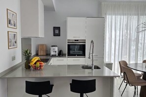 A modern kitchen with white cabinets, a built-in oven, and a coffee maker. There is a fruit bowl on the counter and a dining area with a table and chairs. - Villa LUISA La Marina Alicante with private pool 3 bedrooms & 3 bathrooms (VC)