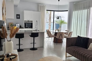 A modern kitchen with white cabinets, a dining area with a round table and chairs, and a living area with a sofa and a coffee table. - Villa LUISA La Marina Alicante with private pool 3 bedrooms & 3 bathrooms (VC)