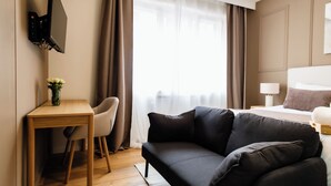 Une chambre d’hôtel avec un bureau en bois, un canapé noir, un lit avec une tête de lit blanche et une grande fenêtre ornée de rideaux légers.