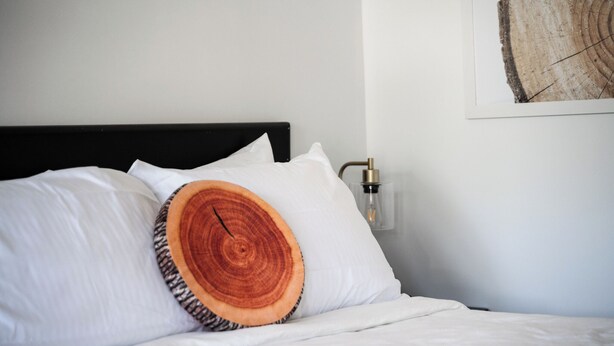 Een bed met wit beddengoed, een kussen in de vorm van een houten boomring en een wandlampje.