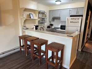 A kitchen with a central island, wooden stools, a refrigerator with magnets, and a microwave. - Modern 1BR Snowcrest Condo: 5-Min Walk to Soaring Eagle Lift (Snowshoe)