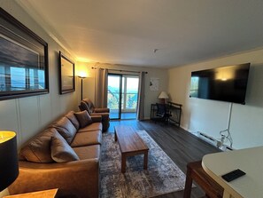 A living room with a sofa, a coffee table, a TV, and a view through a glass door. - Modern 1BR Snowcrest Condo: 5-Min Walk to Soaring Eagle Lift (Snowshoe)