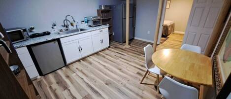 Cocina pequeña con armarios blancos, microondas y lavaplatos. Hay una zona de comedor con una mesa de madera y dos sillas.