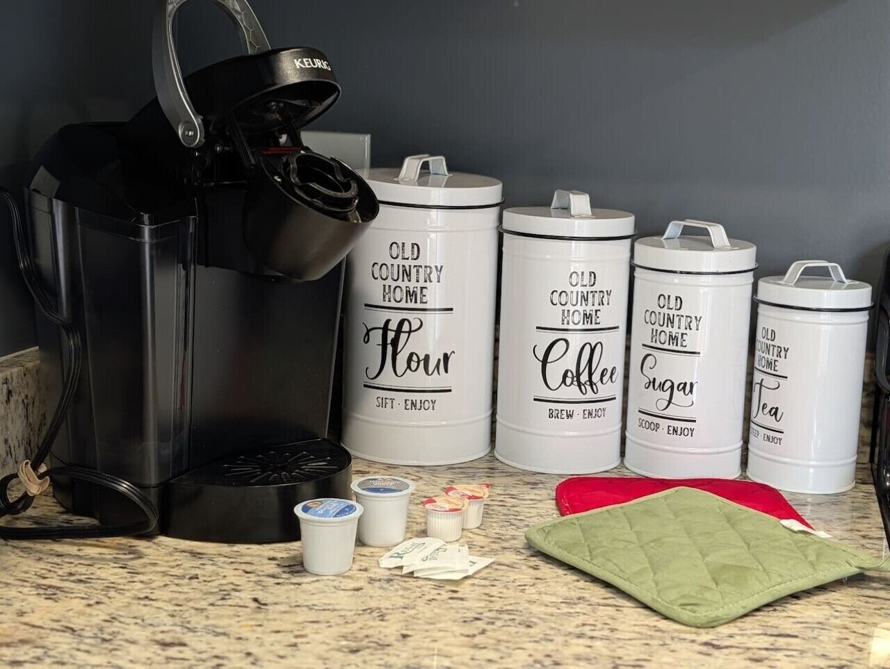 Une machine à café, quatre récipients blancs étiquetés avec des indications sur leur contenu, et quelques petits objets posés sur un plan de travail.