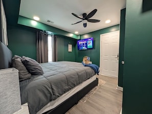 Un dormitorio con cama, ventilador de techo, televisor en la pared y una puerta.